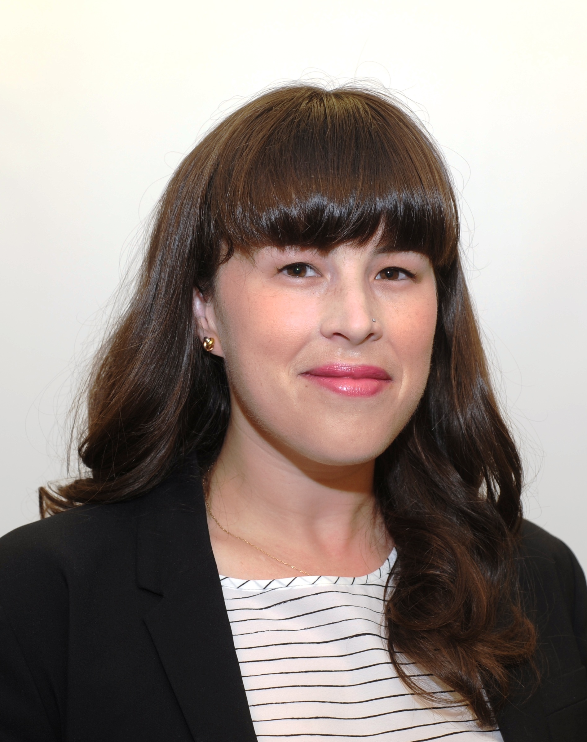Head-and-shoulders portrait of a person with long dark hair and bangs, wearing a black blazer and a white striped top, photographed against a plain light-colored background